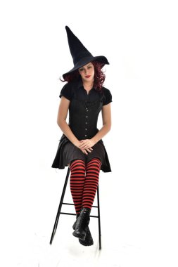 tam uzunlukta uzun siyah pelerin, sivri şapka ve cadı kostümü giyen kırmızı saçlı kız portresi. beyaz stüdyo arka plan üzerinde izole sandalyeye oturmuş poz.