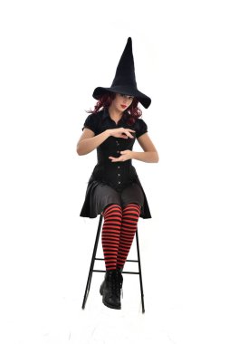 tam uzunlukta uzun siyah pelerin, sivri şapka ve cadı kostümü giyen kırmızı saçlı kız portresi. beyaz stüdyo arka plan üzerinde izole sandalyeye oturmuş poz.
