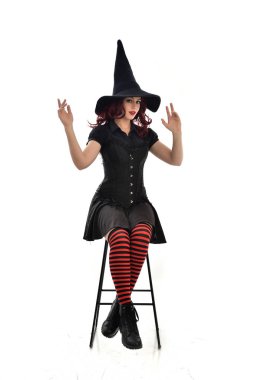 tam uzunlukta uzun siyah pelerin, sivri şapka ve cadı kostümü giyen kırmızı saçlı kız portresi. beyaz stüdyo arka plan üzerinde izole sandalyeye oturmuş poz.