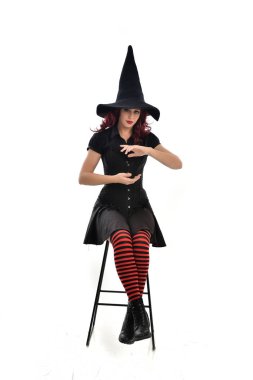 tam uzunlukta uzun siyah pelerin, sivri şapka ve cadı kostümü giyen kırmızı saçlı kız portresi. beyaz stüdyo arka plan üzerinde izole sandalyeye oturmuş poz.