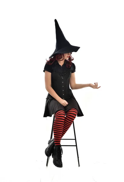 長い黒マント とんがり帽子を被った 魔女の衣装を身に着けている赤髪の女の子の完全な長さの肖像画 ホワイト スタジオの背景に分離されて椅子に座りポーズ — ストック写真