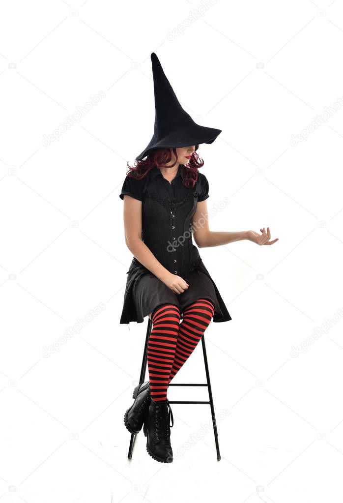 full length portrait of red haired girl wearing long black cloak, pointy hat and witch costume. seated pose on chair, isolated on white studio background.