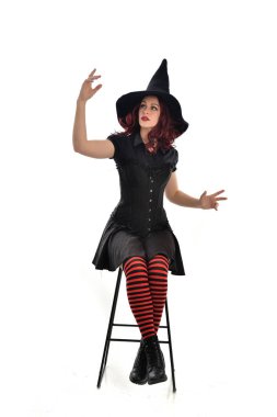 tam uzunlukta uzun siyah pelerin, sivri şapka ve cadı kostümü giyen kırmızı saçlı kız portresi. beyaz stüdyo arka plan üzerinde izole sandalyeye oturmuş poz.