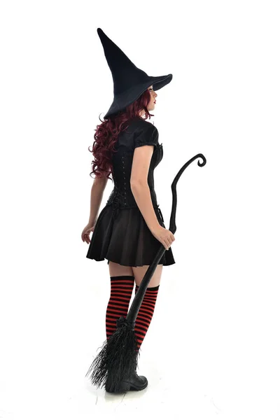 ほうきを持っている黒魔女衣装と先のとがった帽子を身に着けている赤髪少女の完全な長さの肖像画 立ちポーズ 白いスタジオ背景に分離 ストック写真 C Faestock