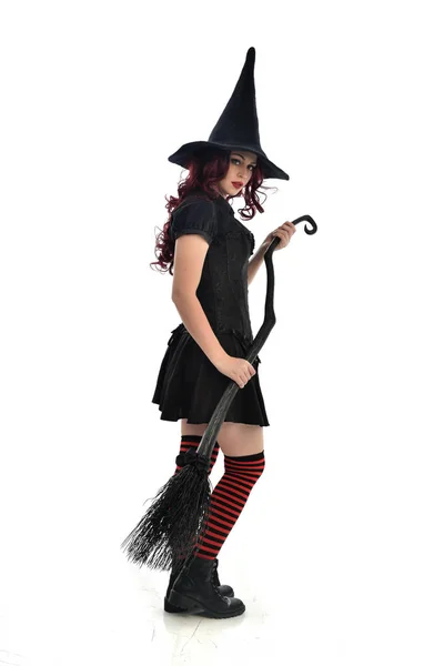 ほうきを持っている黒魔女衣装と先のとがった帽子を身に着けている赤髪少女の完全な長さの肖像画 立ちポーズ 白いスタジオ背景に分離 — ストック写真