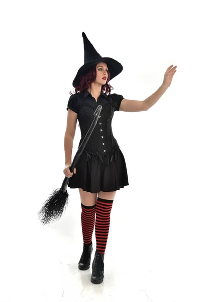 ほうきを持っている黒魔女衣装と先のとがった帽子を身に着けている赤髪少女の完全な長さの肖像画 立ちポーズ 白いスタジオ背景に分離 — ストック写真