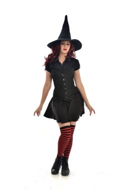 tam uzunlukta portresi kırmızı saçlı kız kara cadı kostüm ve sivri şapka giyiyor. ayakta poz, beyaz stüdyo arka plan üzerinde izole.