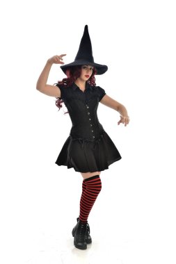 tam uzunlukta portresi kırmızı saçlı kız kara cadı kostüm ve sivri şapka giyiyor. ayakta poz, beyaz stüdyo arka plan üzerinde izole.