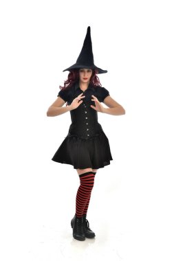 tam uzunlukta portresi kırmızı saçlı kız kara cadı kostüm ve sivri şapka giyiyor. ayakta poz, beyaz stüdyo arka plan üzerinde izole.