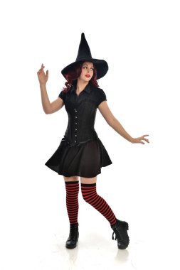 tam uzunlukta portresi kırmızı saçlı kız kara cadı kostüm ve sivri şapka giyiyor. ayakta poz, beyaz stüdyo arka plan üzerinde izole.