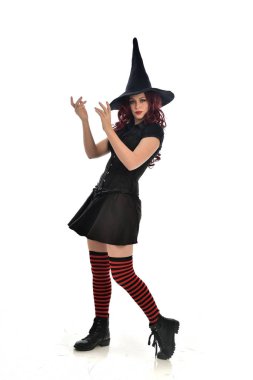 tam uzunlukta portresi kırmızı saçlı kız kara cadı kostüm ve sivri şapka giyiyor. ayakta poz, beyaz stüdyo arka plan üzerinde izole.