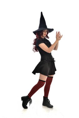 tam uzunlukta portresi kırmızı saçlı kız kara cadı kostüm ve sivri şapka giyiyor. ayakta poz, beyaz stüdyo arka plan üzerinde izole.