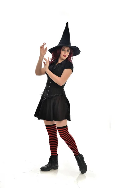 黒魔女の衣装と先のとがった帽子を身に着けている赤い髪の女の子の完全な長さの肖像画 立ちポーズ 白いスタジオ背景に分離 — ストック写真