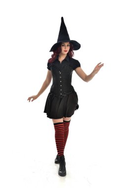 tam uzunlukta portresi kırmızı saçlı kız kara cadı kostüm ve sivri şapka giyiyor. ayakta poz, beyaz stüdyo arka plan üzerinde izole.