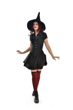 tam uzunlukta portresi kırmızı saçlı kız kara cadı kostüm ve sivri şapka giyiyor. ayakta poz, beyaz stüdyo arka plan üzerinde izole.