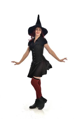 tam uzunlukta portresi kırmızı saçlı kız kara cadı kostüm ve sivri şapka giyiyor. ayakta poz, beyaz stüdyo arka plan üzerinde izole.
