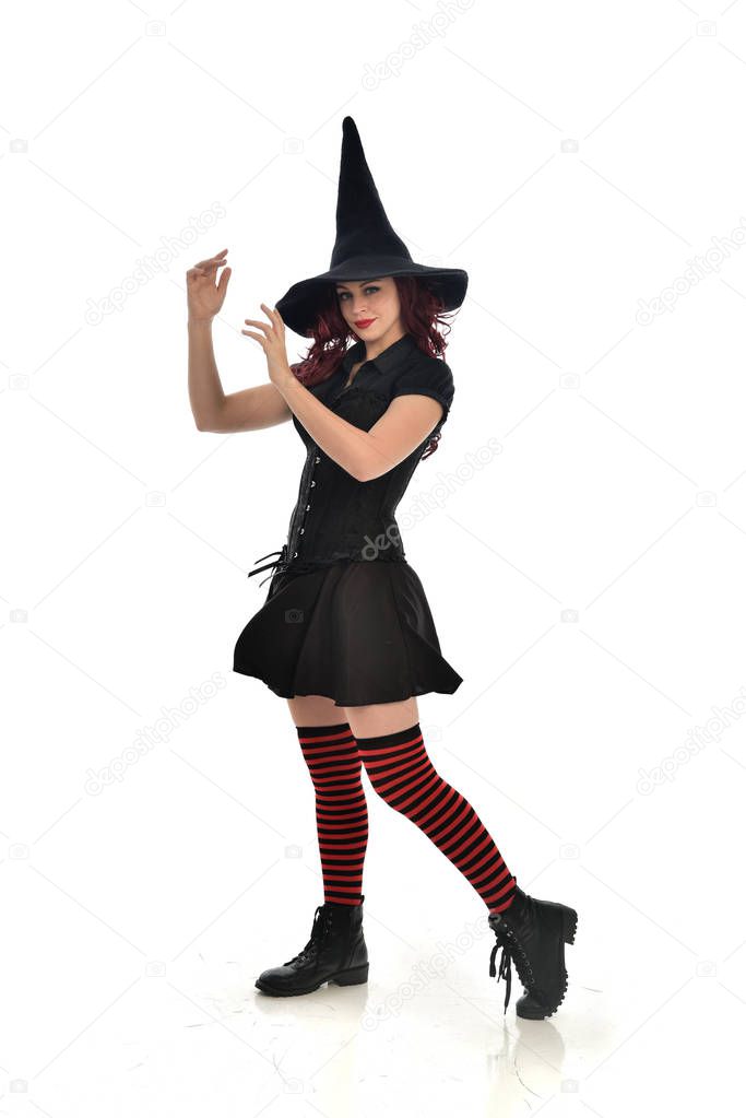 full length portrait of red haired girl wearing black witch costume and pointy hat.  standing pose, isolated on white studio background.