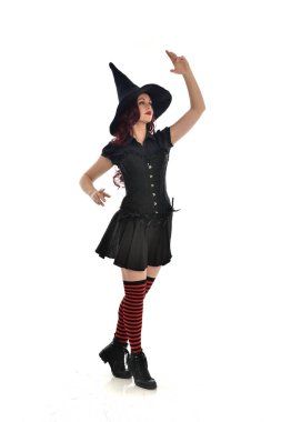 tam uzunlukta portresi kırmızı saçlı kız kara cadı kostüm ve sivri şapka giyiyor. ayakta poz, beyaz stüdyo arka plan üzerinde izole.
