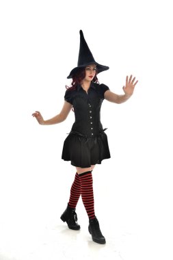 tam uzunlukta portresi kırmızı saçlı kız kara cadı kostüm ve sivri şapka giyiyor. ayakta poz, beyaz stüdyo arka plan üzerinde izole.
