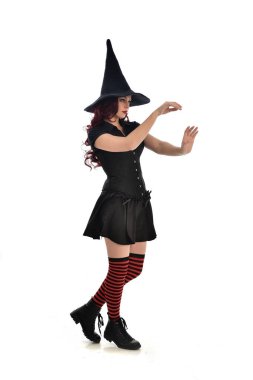 tam uzunlukta portresi kırmızı saçlı kız kara cadı kostüm ve sivri şapka giyiyor. ayakta poz, beyaz stüdyo arka plan üzerinde izole.