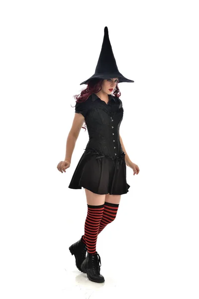 黒魔女の衣装と先のとがった帽子を身に着けている赤い髪の女の子の完全な長さの肖像画 立ちポーズ 白いスタジオ背景に分離 — ストック写真