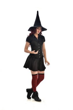 tam uzunlukta portresi kırmızı saçlı kız kara cadı kostüm ve sivri şapka giyiyor. ayakta poz, beyaz stüdyo arka plan üzerinde izole.