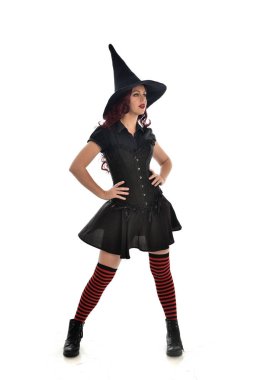 tam uzunlukta portresi kırmızı saçlı kız kara cadı kostüm ve sivri şapka giyiyor. ayakta poz, beyaz stüdyo arka plan üzerinde izole.