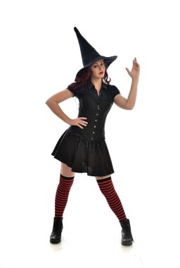tam uzunlukta portresi kırmızı saçlı kız kara cadı kostüm ve sivri şapka giyiyor. ayakta poz, beyaz stüdyo arka plan üzerinde izole.