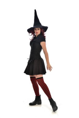 tam uzunlukta portresi kırmızı saçlı kız kara cadı kostüm ve sivri şapka giyiyor. ayakta poz, beyaz stüdyo arka plan üzerinde izole.