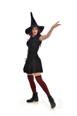 tam uzunlukta portresi kırmızı saçlı kız kara cadı kostüm ve sivri şapka giyiyor. ayakta poz, beyaz stüdyo arka plan üzerinde izole.