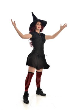 tam uzunlukta portresi kırmızı saçlı kız kara cadı kostüm ve sivri şapka giyiyor. ayakta poz, beyaz stüdyo arka plan üzerinde izole.