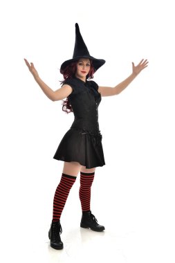 tam uzunlukta portresi kırmızı saçlı kız kara cadı kostüm ve sivri şapka giyiyor. ayakta poz, beyaz stüdyo arka plan üzerinde izole.