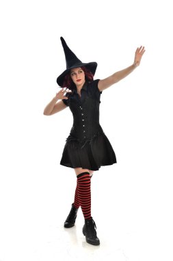 tam uzunlukta portresi kırmızı saçlı kız kara cadı kostüm ve sivri şapka giyiyor. ayakta poz, beyaz stüdyo arka plan üzerinde izole.