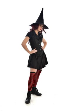 tam uzunlukta portresi kırmızı saçlı kız kara cadı kostüm ve sivri şapka giyiyor. ayakta poz, beyaz stüdyo arka plan üzerinde izole.