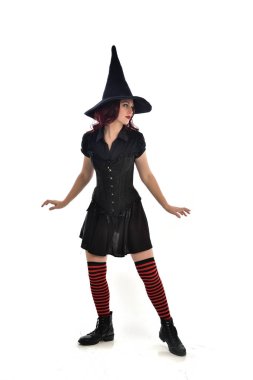 tam uzunlukta portresi kırmızı saçlı kız kara cadı kostüm ve sivri şapka giyiyor. ayakta poz, beyaz stüdyo arka plan üzerinde izole.