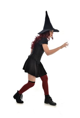 tam uzunlukta portresi kırmızı saçlı kız kara cadı kostüm ve sivri şapka giyiyor. ayakta poz, beyaz stüdyo arka plan üzerinde izole.
