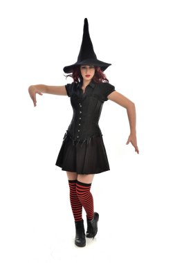 tam uzunlukta portresi kırmızı saçlı kız kara cadı kostüm ve sivri şapka giyiyor. ayakta poz, beyaz stüdyo arka plan üzerinde izole.