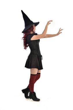 tam uzunlukta portresi kırmızı saçlı kız kara cadı kostüm ve sivri şapka giyiyor. ayakta poz, beyaz stüdyo arka plan üzerinde izole.