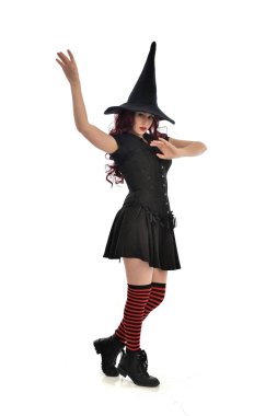 tam uzunlukta portresi kırmızı saçlı kız kara cadı kostüm ve sivri şapka giyiyor. ayakta poz, beyaz stüdyo arka plan üzerinde izole.