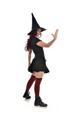 tam uzunlukta portresi kırmızı saçlı kız kara cadı kostüm ve sivri şapka giyiyor. arka beyaz stüdyo arka plan üzerinde izole kamera ile ayakta poz.