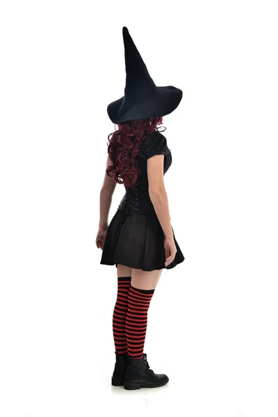 黒魔女の衣装と先のとがった帽子を身に着けている赤い髪の女の子の完全な長さの肖像画 ホワイト スタジオの背景に分離カメラに背中に立ちポーズ — ストック写真