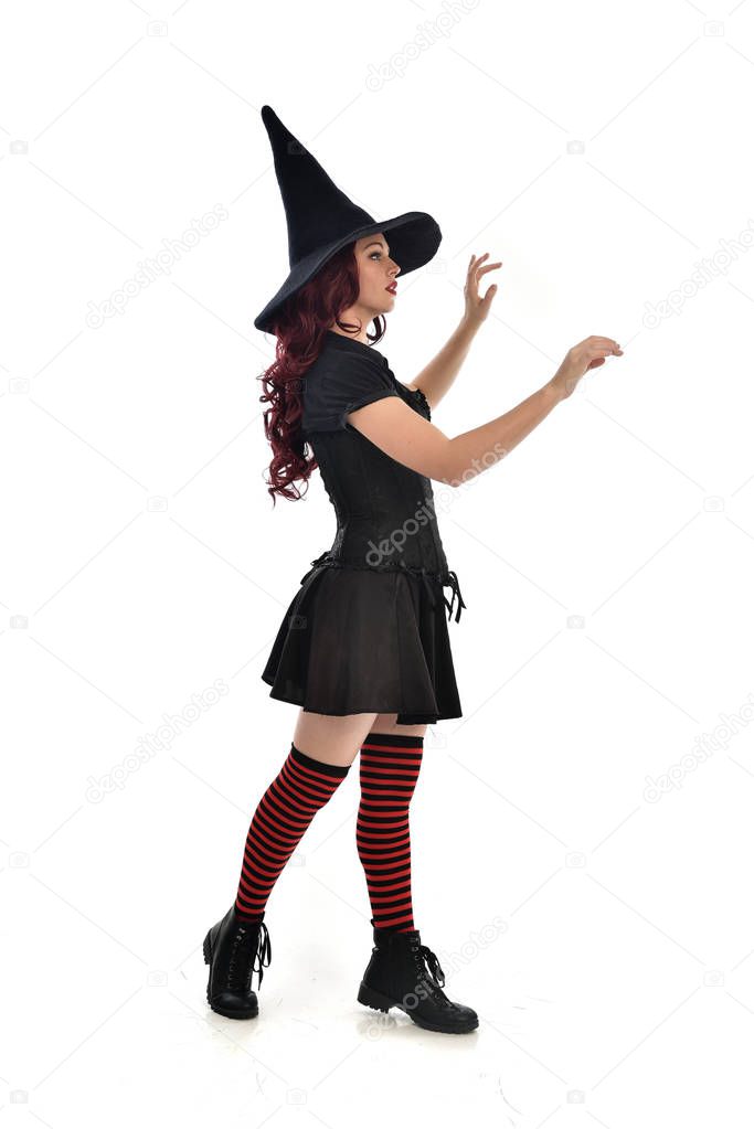 full length portrait of red haired girl wearing black witch costume and pointy hat.  standing pose, isolated on white studio background.