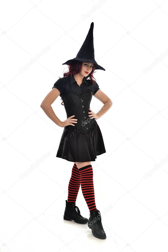 full length portrait of red haired girl wearing black witch costume and pointy hat.  standing pose, isolated on white studio background.
