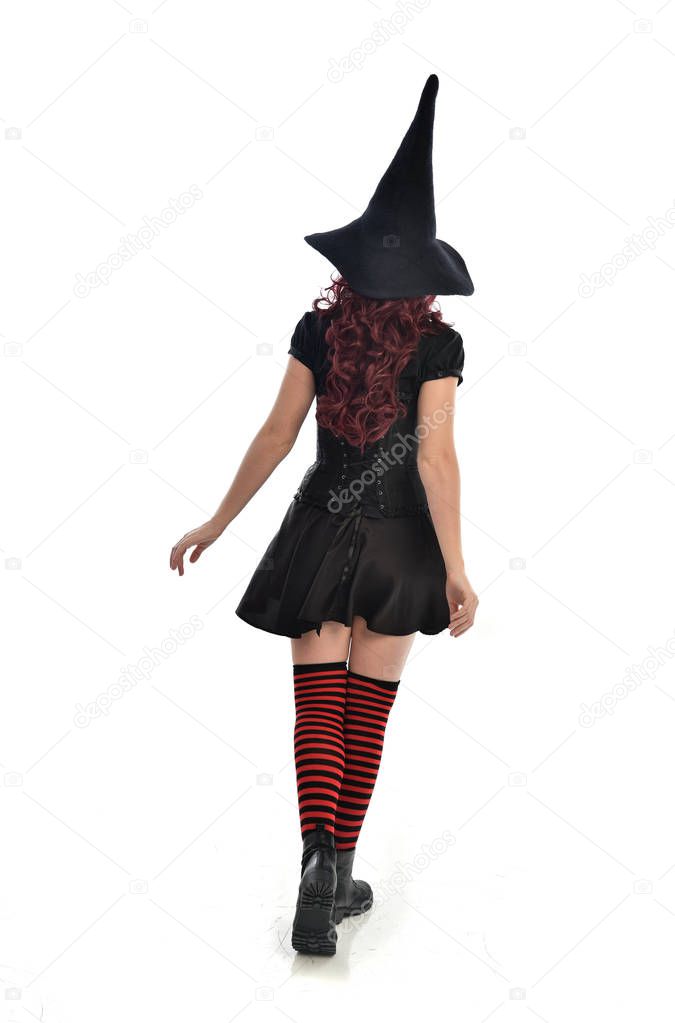 full length portrait of red haired girl wearing black witch costume and pointy hat.  standing pose with back to the camera, isolated on white studio background.