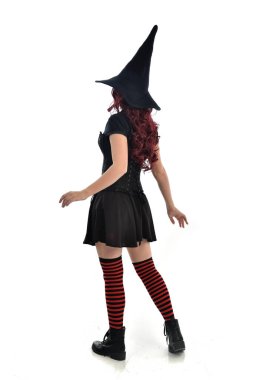 tam uzunlukta portresi kırmızı saçlı kız kara cadı kostüm ve sivri şapka giyiyor. arka beyaz stüdyo arka plan üzerinde izole kamera ile ayakta poz.