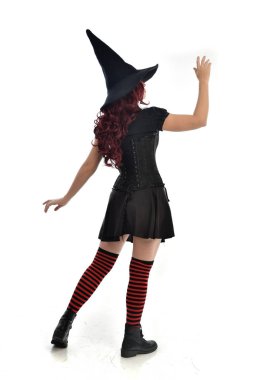 tam uzunlukta portresi kırmızı saçlı kız kara cadı kostüm ve sivri şapka giyiyor. arka beyaz stüdyo arka plan üzerinde izole kamera ile ayakta poz.