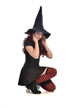 tam uzunlukta portresi kırmızı saçlı kız kara cadı kostüm ve sivri şapka giyiyor. beyaz stüdyo arka plan üzerinde izole oturmuş poz.