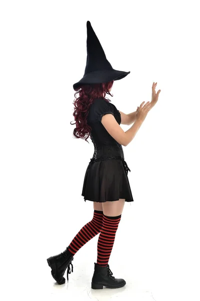 黒魔女の衣装と先のとがった帽子を身に着けている赤い髪の女の子の完全な長さの肖像画 ホワイト スタジオの背景に分離カメラに背中に立ちポーズ — ストック写真