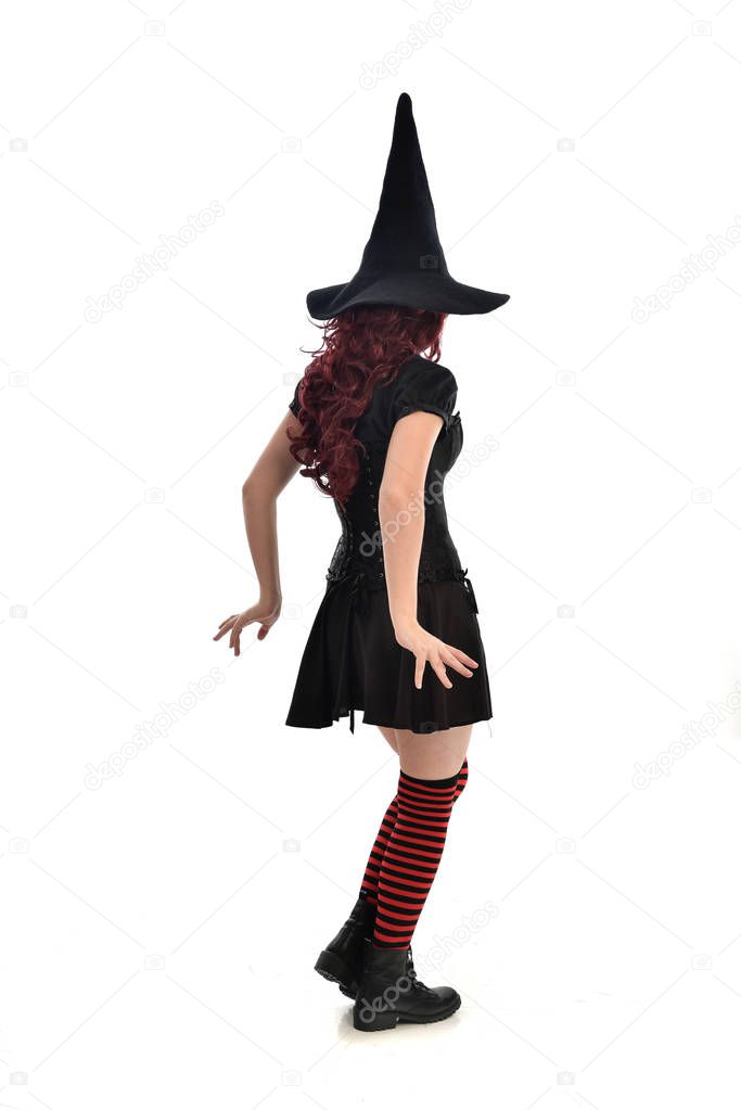 full length portrait of red haired girl wearing black witch costume and pointy hat.  standing pose with back to the camera, isolated on white studio background.