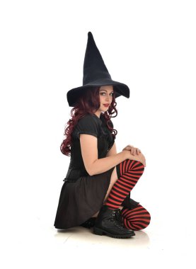 tam uzunlukta portresi kırmızı saçlı kız kara cadı kostüm ve sivri şapka giyiyor. beyaz stüdyo arka plan üzerinde izole oturmuş poz.